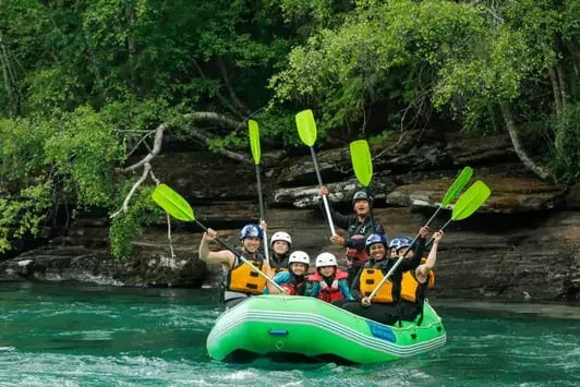 Menschen, die ein Rafting-Abenteuer mit der Familie auf einem klaren, türkisfarbenen Fluss genießen, umgeben von üppigem, grünem Wald, ihre Paddel heben und lächeln